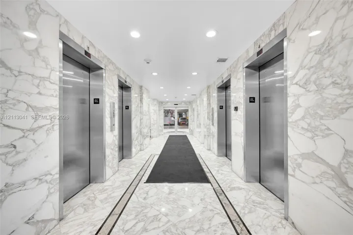 Elevator lobby featuring floor-to-ceiling stone wall cladding, polished stone flooring with decorative inlay, stainless steel elevator doors, and recessed ceiling lighting