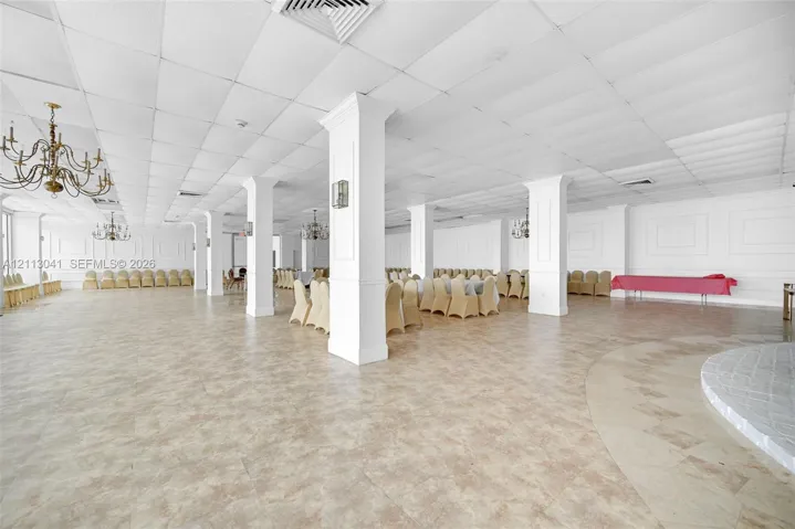 Expansive open-concept hall featuring a dropped acoustic tile ceiling, structural columns with wainscoting detail, wall-mounted light fixtures, tile flooring, and multiple ornate chandeliers
