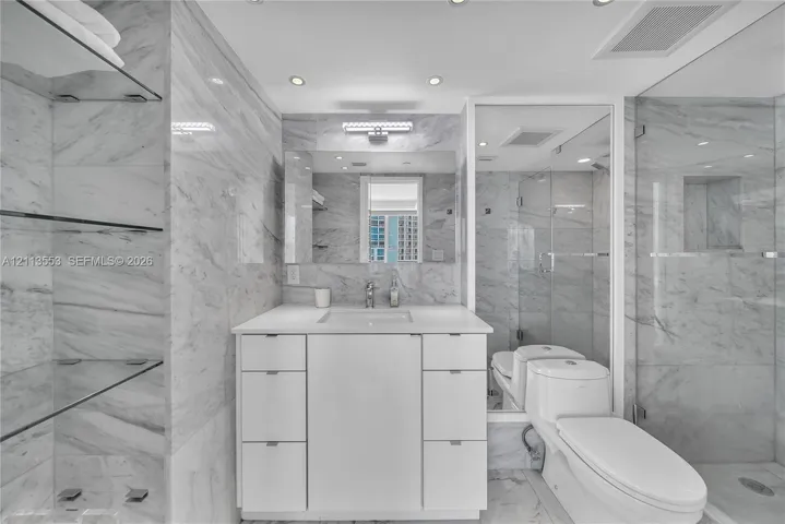 Bathroom featuring floor-to-ceiling stone-tiled walls, a white vanity with integrated basin, recessed lighting, glass shelving, and a frameless glass-enclosed shower