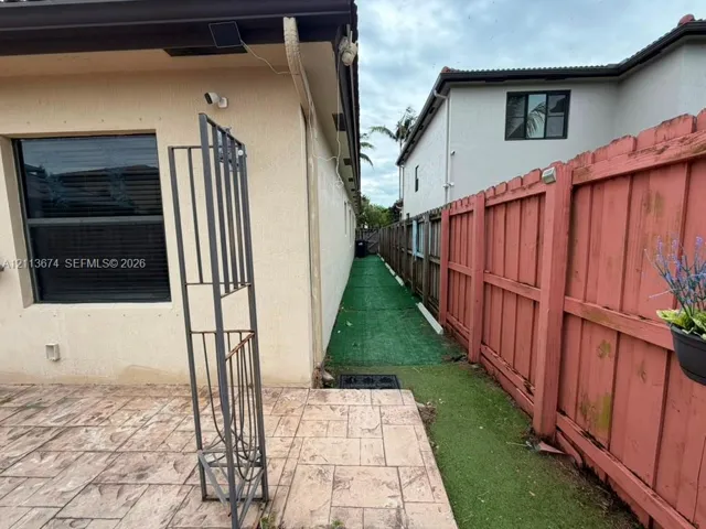 Stucco exterior wall featuring a window with dark-tinted glass, finished concrete patio, and side yard with synthetic turf and a wood-slat privacy fence