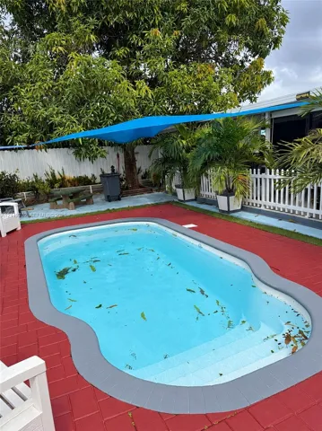 In-ground pool with molded steps, surrounded by red brick-patterned paving and a gray bullnose coping border, complemented by white picket fencing and a blue overhead tension shade sail