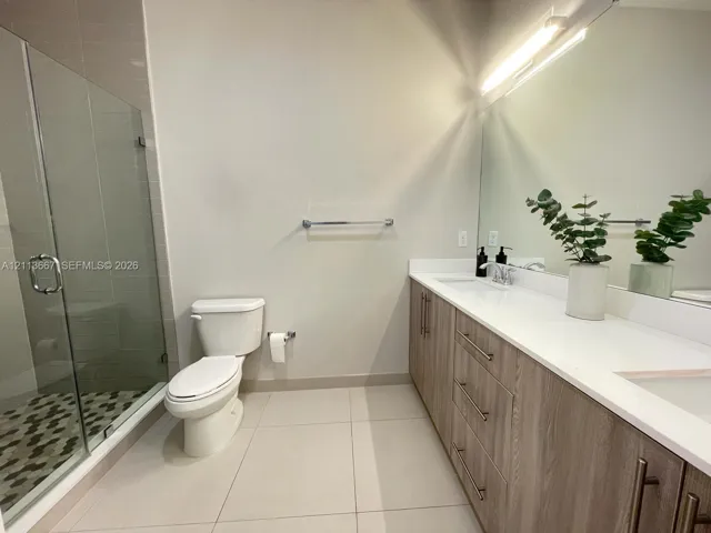 Bathroom featuring a wood-finish double vanity with white countertop, integrated dual sinks, frameless glass-enclosed shower with mosaic tile flooring, and a full-width vanity mirror