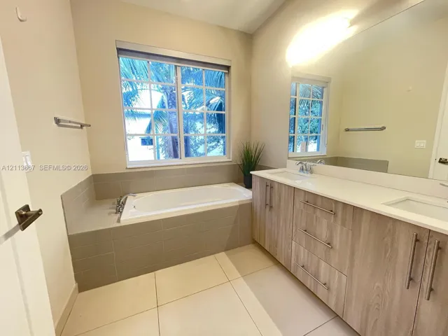 Bathroom featuring a dual-sink vanity with wood-finish cabinetry, a drop-in soaking tub with a tiled surround, a large wall-mounted mirror, and tile flooring