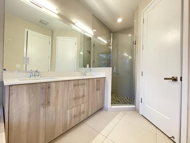 Dual-vanity cabinetry with wood-finish doors, solid-surface countertops, full-width mirror, and a glass-enclosed shower with tiled flooring