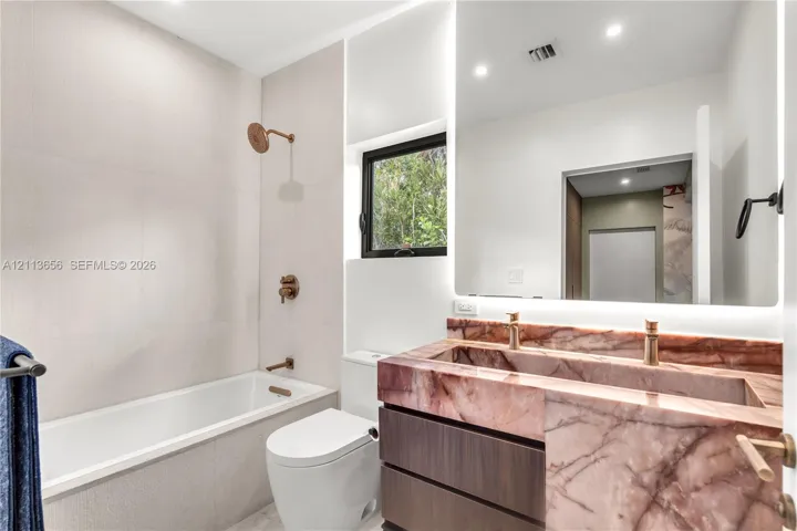 Bathroom featuring a custom integrated sink with stone vanity, wood-finish cabinetry, wall-to-wall LED-backlit mirror, soaking tub, and metallic fixtures