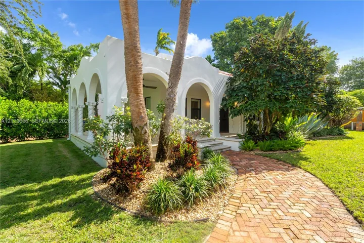 Spanish Colonial exterior featuring a covered arched portico, decorative parapet roofline, brick paver walkway, and mature professional landscaping