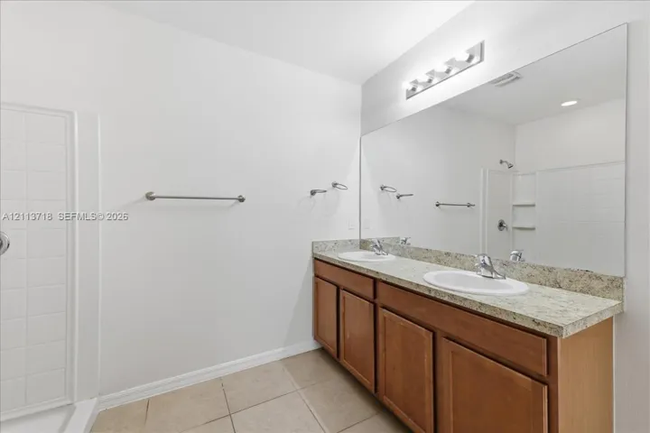 Dual-sink vanity with stone-finish countertop, full-width mirror, tile flooring, and integrated shower enclosure