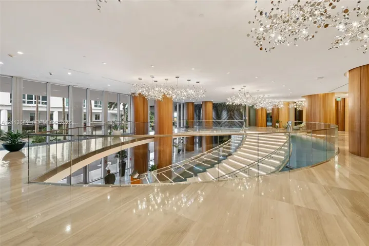 Grand atrium featuring polished stone flooring, custom architectural wood-finish columns, a sweeping curved staircase with glass railing, and oversized decorative chandeliers