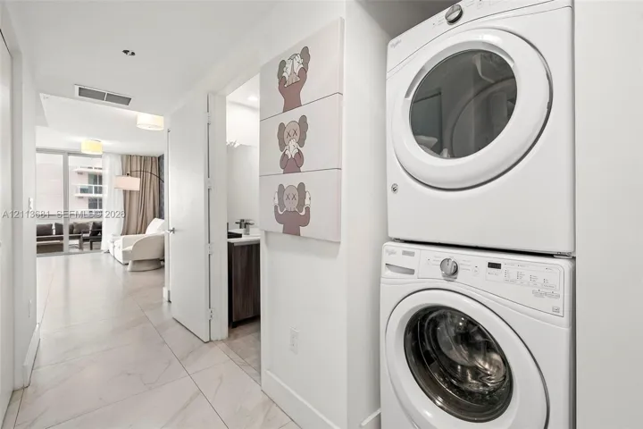 Stacked front-load laundry unit featuring a white finish and large-format porcelain tile flooring