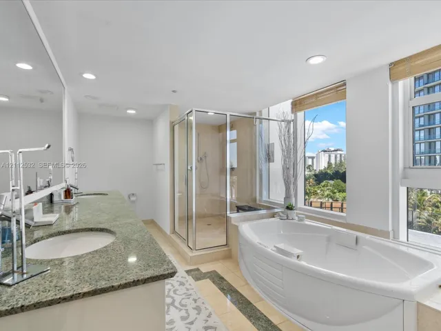 Dual-sink vanity with natural stone countertop, corner glass-enclosed shower, deep soaking tub, tile flooring with stone inlay, and large windows with integrated blinds
