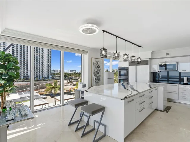 Kitchen featuring high-gloss cabinetry, stone-topped center island with waterfall edge, built-in double wall ovens, five-light linear pendant fixture, and floor-to-ceiling windows