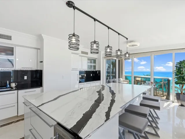 Kitchen featuring a large central island with veined stone countertop, high-gloss white cabinetry, black tile backsplash, and linear pendant lighting, framed by floor-to-ceiling glass doors with ocean scenery