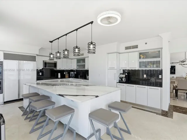 Kitchen featuring a large waterfall-edge island, high-gloss white cabinetry, contrasting dark stone countertops, integrated stainless steel appliances, and linear industrial-style pendant lighting