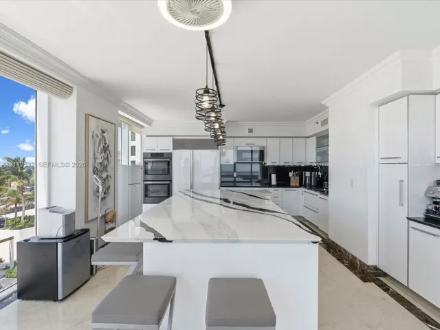 Kitchen featuring a substantial waterfall-edge island with stone countertop, flat-panel white cabinetry, built-in stainless steel appliances, multi-pendant track lighting, and light-toned tile flooring