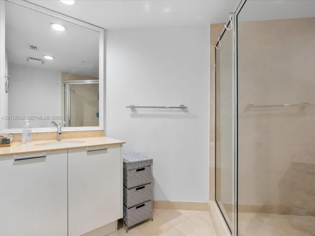 Bathroom vanity with a light-toned countertop, integrated sink, and white cabinetry, featuring a large framed mirror, stone-tile flooring, and a walk-in shower enclosure with a glass door