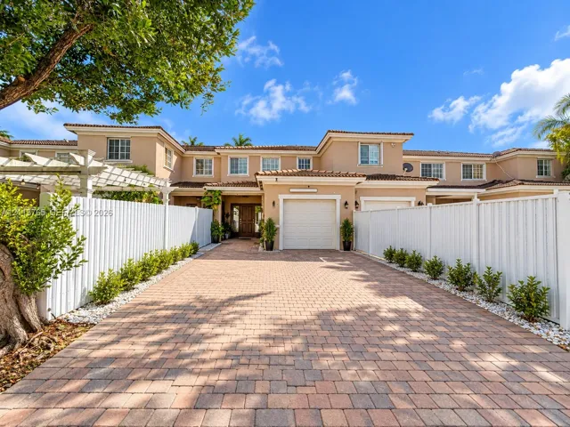 Stucco exterior with a multi-gabled tile roof, paved driveway, attached garage, and white vinyl perimeter fencing
