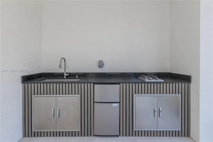 Outdoor kitchen featuring stone-finish countertops, stainless steel sink with pull-down faucet, built-in cooktop, under-counter refrigeration, and slatted wood-finish cabinetry