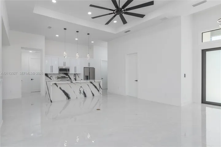 Open-concept interior featuring high tray ceilings with a multi-blade fan, polished light-toned flooring, a waterfall-edge kitchen island, glass pendant lighting, and sleek cabinetry