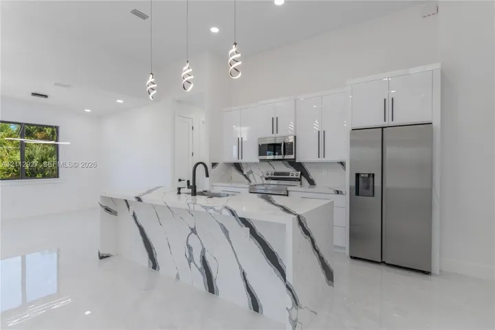 Modern kitchen featuring high-gloss cabinetry, waterfall edge island with stone-finish countertop, stainless steel appliances, matte black plumbing fixtures, and contemporary pendant lighting