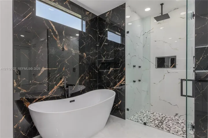 Freestanding soaking tub, walk-in glass-enclosed shower with a rainfall showerhead, black and white veined tile wall finishes, recessed lighting, and built-in shower niche