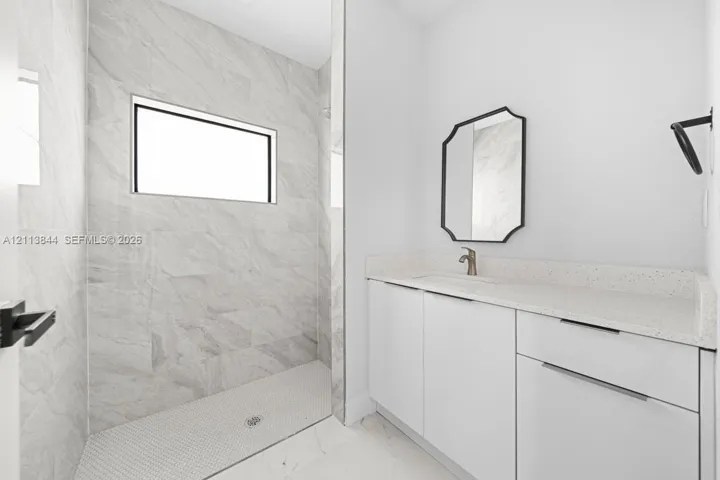Walk-in shower featuring large-format stone-tile surround, light-colored quartz vanity countertop, white flat-panel cabinetry, hexagonal floor tiling, and dark-finish hardware