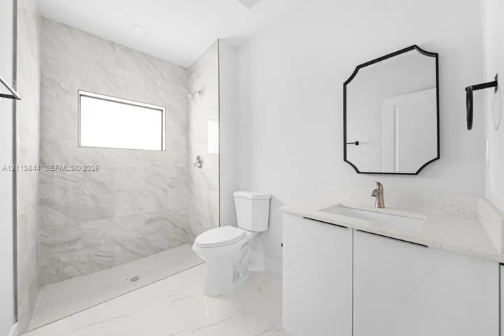 Full bathroom featuring a walk-in shower with stone-finish tile surround, white vanity cabinet with quartz countertop, undermount sink, and geometric wall-mounted mirror