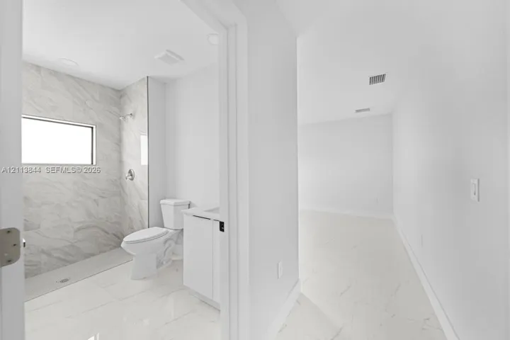 Bathroom featuring large-format light-toned tile flooring, walk-in shower with stone-style wall surround, transom window, and white cabinetry