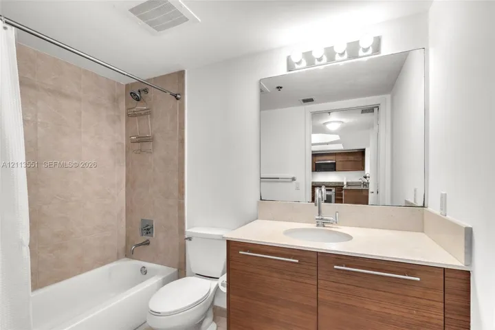 Bathroom featuring a wood-finish vanity with a light-toned solid surface countertop, rectangular mirror, light-bar fixture, and tub-shower combination with textured surround tiling
