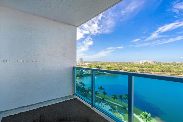 Private balcony featuring a textured concrete ceiling and walls with a blue-tinted glass and metal railing system, overlooking a body of water and natural landscape