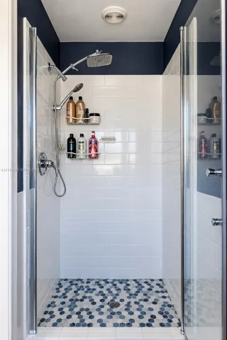 Walk-in shower enclosure featuring white subway tile walls, geometric hexagonal floor tile, chrome rain showerhead with a secondary handheld sprayer, and glass panel doors