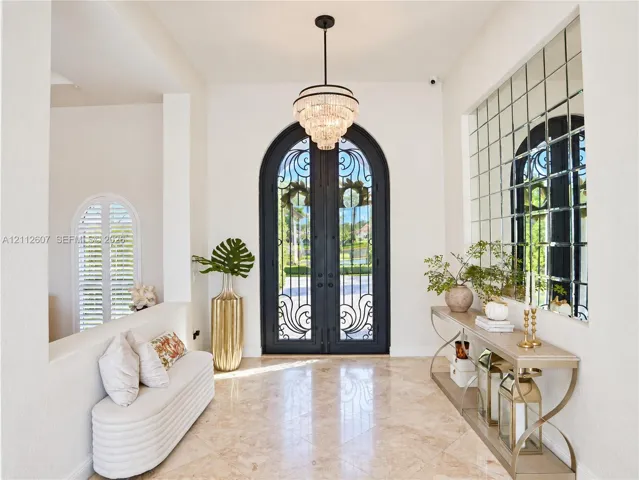 Foyer featuring arched double iron entry doors, light-toned stone flooring, a multi-tiered crystal chandelier, and an expansive grid-pattern wall mirror