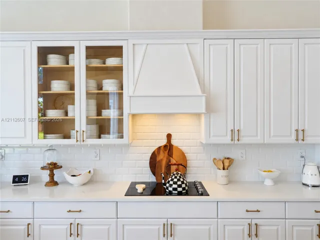 Kitchen featuring white cabinetry with glass-front uppers, a custom range hood, subway tile backsplash, solid-surface countertops, and gold-tone hardware