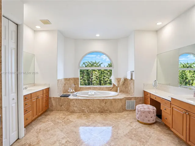Bathroom featuring dual wooden vanities, stone-tile flooring, jetted soaking tub with arched window surround, and large wall-mounted mirrors