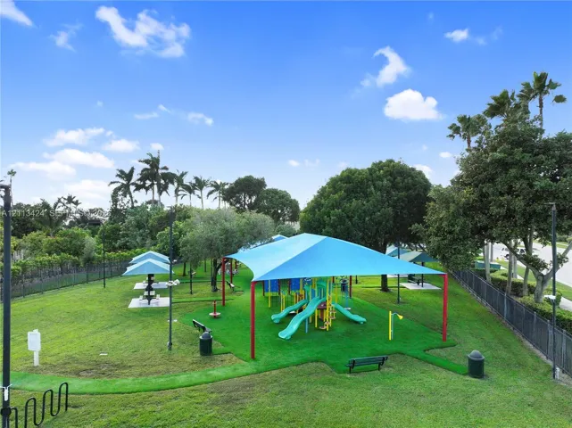 Outdoor recreation area featuring a playground structure with a blue shade canopy, integrated turf surfacing, picnic stations, perimeter fencing, and mature landscaping