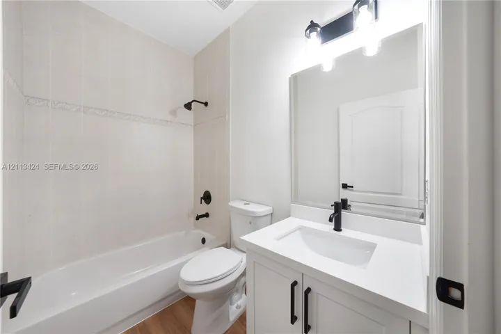 Bathroom featuring a white vanity with a rectangular basin, matte black faucet, full-width vanity mirror, wood-finish flooring, and a tub-shower combination with light-toned tile surround