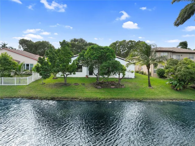 Waterfront property featuring a manicured lawn, mature trees, and a white privacy fence