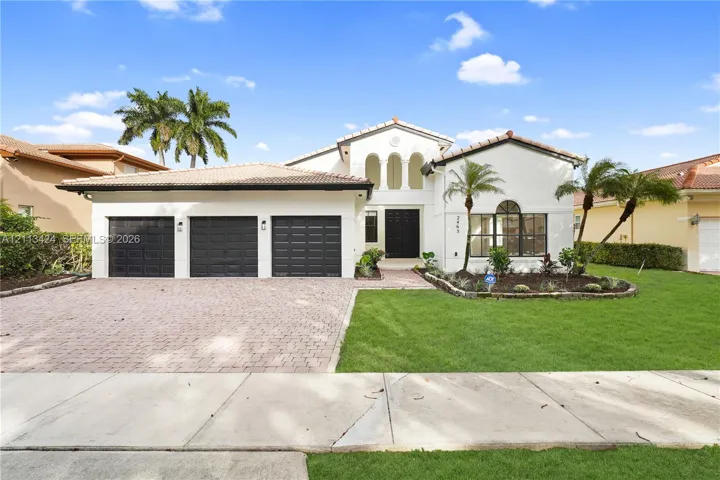 Stucco exterior featuring a tile roof, three-car garage with paneled doors, arched entryway, and a brick-paver driveway (virtually edited out tree)