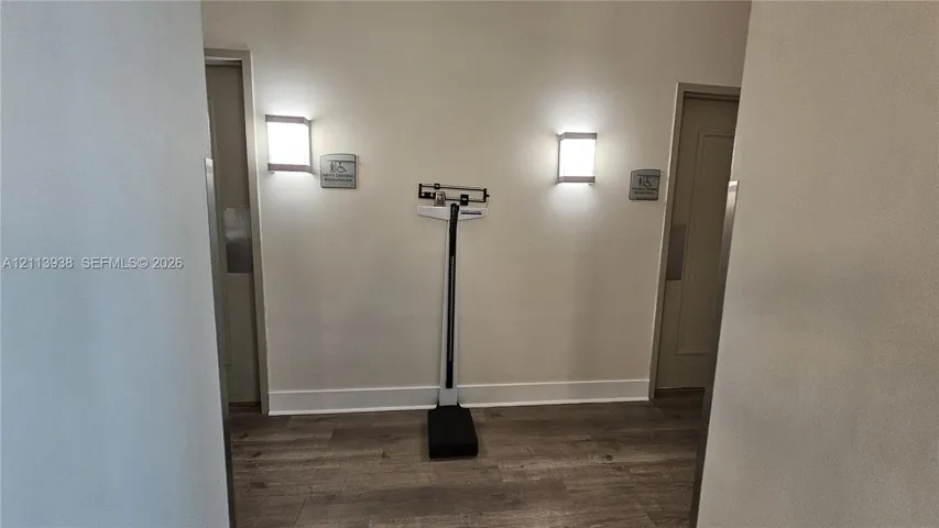 Interior hall featuring wood-finish flooring, two wall-mounted rectangular light fixtures, and dual entryways with brushed metal door plates