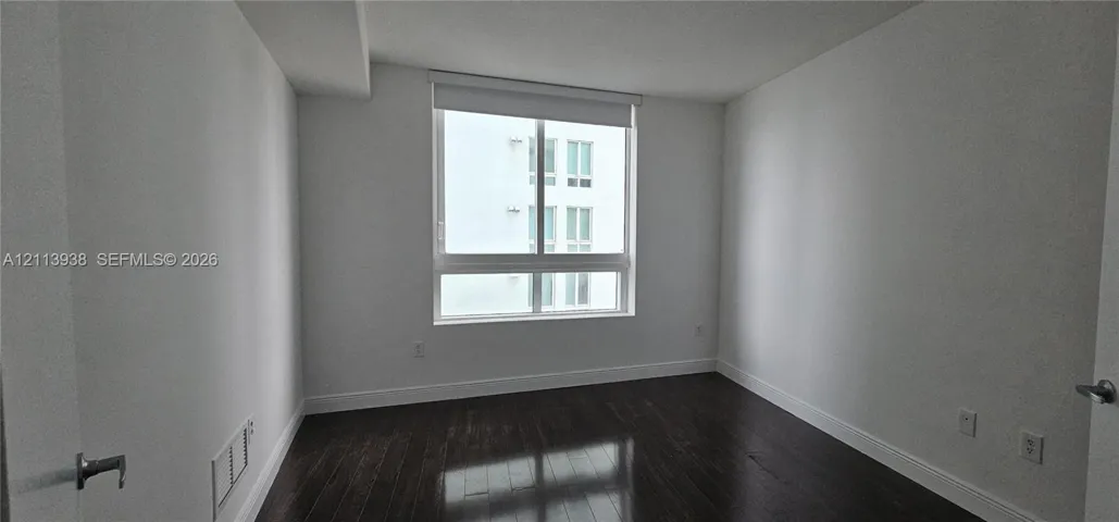 Interior room featuring dark wood-finish flooring, neutral walls, and a large window with a roller shade