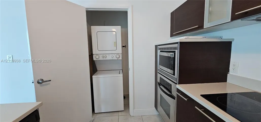 Dark-finish cabinetry, stainless steel wall-mounted oven and microwave, induction cooktop, solid-surface countertops, and built-in stacked laundry unit