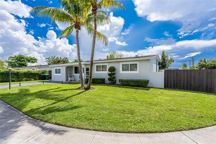 Single-story exterior featuring a light-toned smooth finish, manicured lawn, tall palm trees, and tall wood fencing