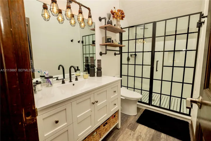 Bathroom vanity featuring white cabinetry, a solid surface countertop, and matte black widespread faucet, complemented by a walk-in shower with a black grid-frame glass enclosure, industrial-style multi-bulb vanity lighting, and wall-mounted pipe-style shelving