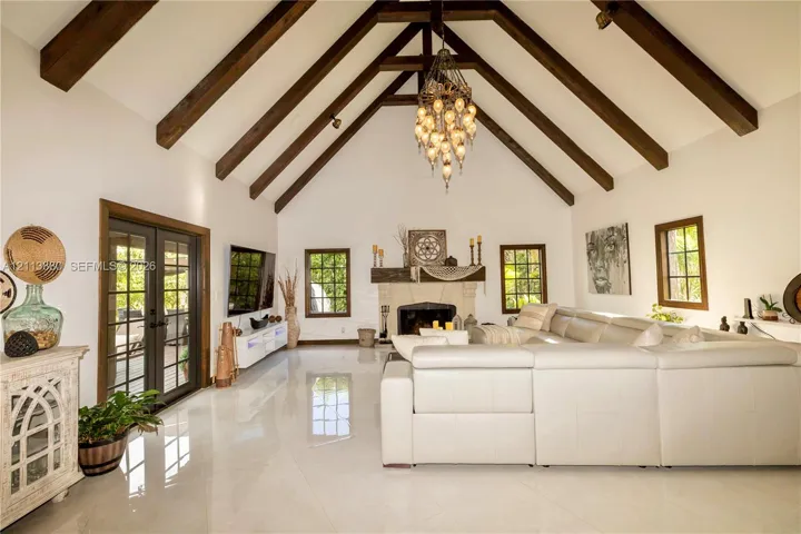 Vaulted ceiling with exposed wood-finish beams, polished tile flooring, stone-surround fireplace with a wooden mantel, and multi-pane glass french doors