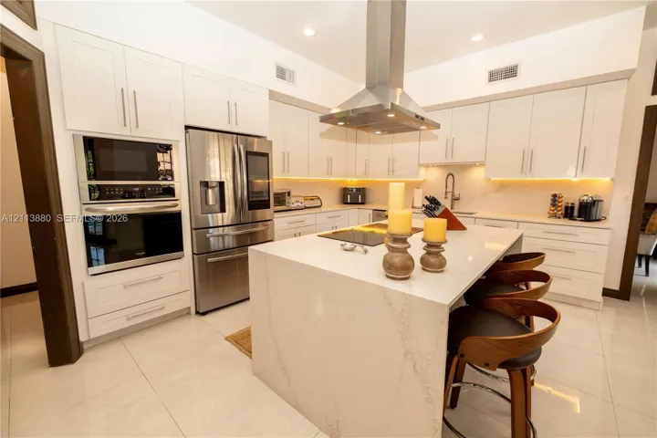 Kitchen featuring white cabinetry, quartz countertops with a waterfall edge island, stainless steel wall-mounted range hood, built-in stainless steel double oven, and integrated cabinet lighting