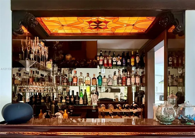 Built-in bar area featuring a stone countertop, mirrored wall backing, multiple glass shelving tiers, and a decorative amber-hued stained glass ceiling inlay framed by carved wood corbels