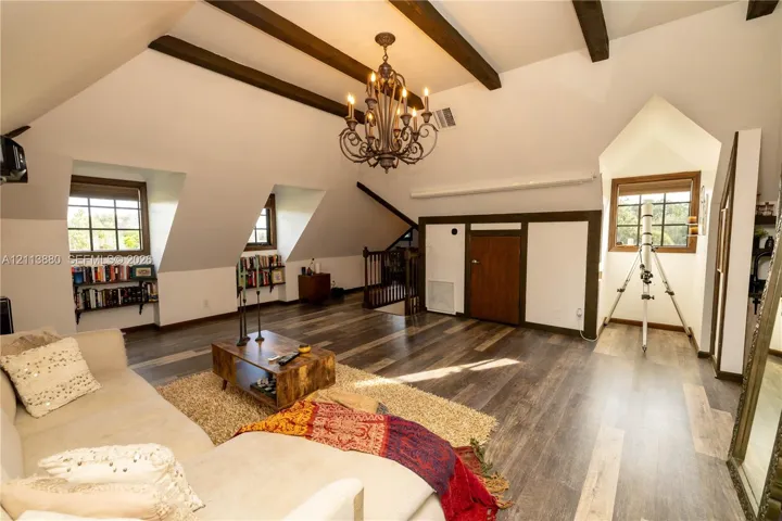 Vaulted ceiling with exposed wood beams, wood-finish flooring, custom built-in shelving, dormer windows, and a multi-arm chandelier