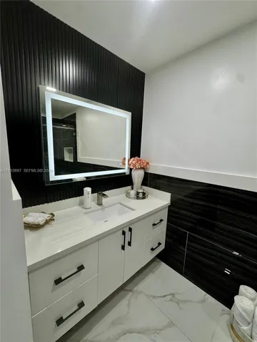 White vanity with clean-lined cabinetry, quartz countertop, integrated rectangular sink, illuminated mirror, and vertical slat accent wall