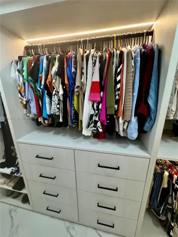 Custom walk-in closet featuring light-toned wood-finish cabinetry, eight soft-close drawers with matte black hardware, integrated linear LED shelf lighting, and stone-look tile flooring