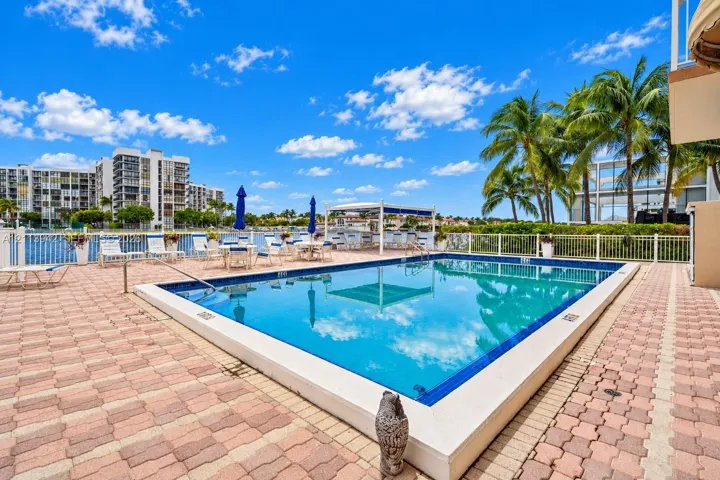 In-ground swimming pool with tile border, brick paver decking, white perimeter fencing, and mature palm trees