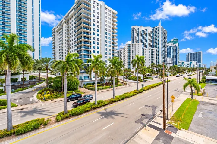 High-rise residential exterior featuring glass balcony railings, white concrete facade, mature palm tree landscaping, and paved multi-lane street access